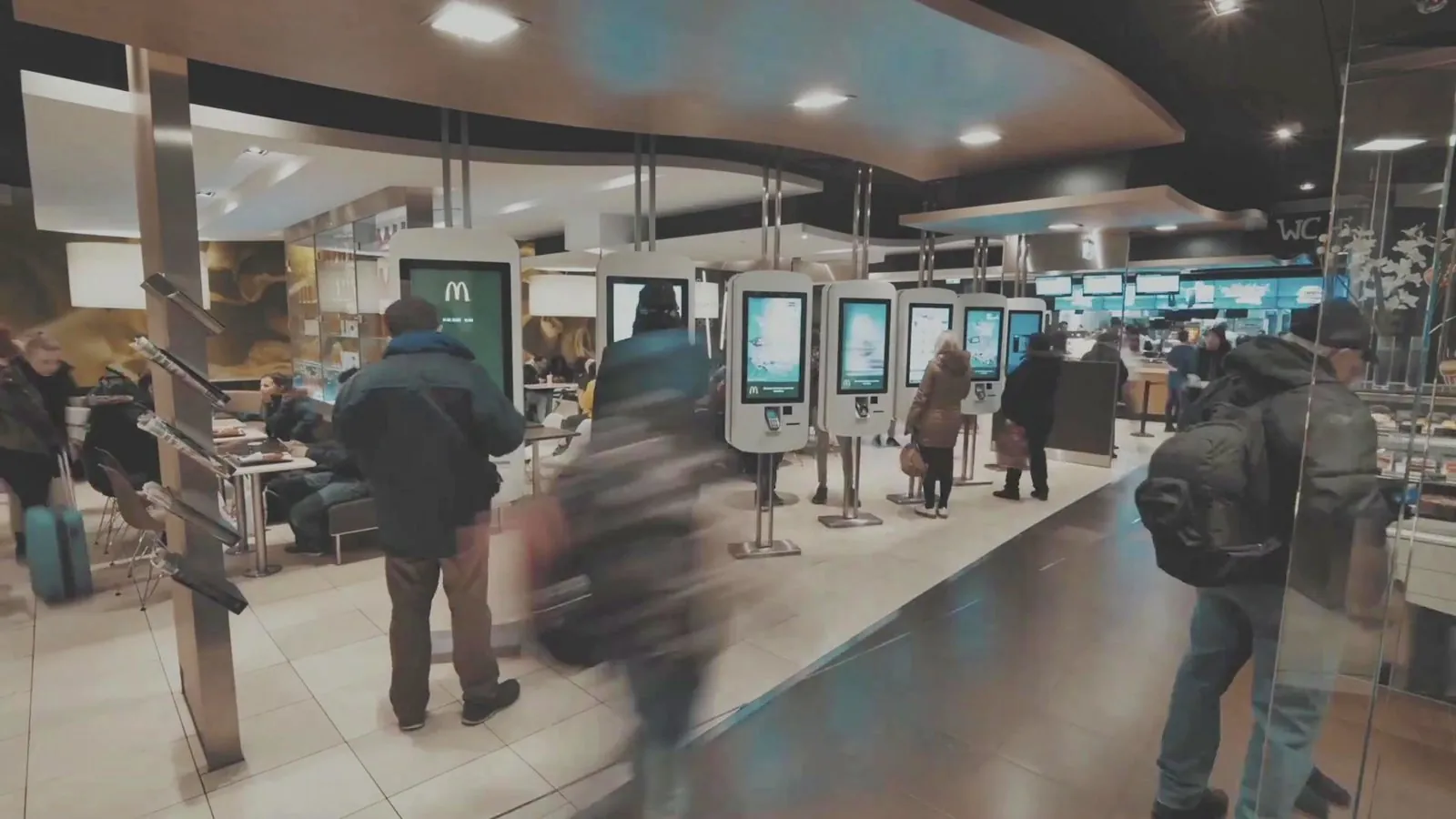 Self-order kiosks in a busy restaurant, with menu boards behind the counter